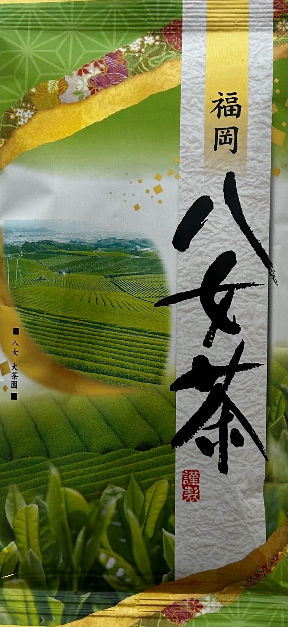 Bildstarke japanische Teetüte, die auf der linken Seite eine kreisrund eingefasste, fotorealistische Landschaftsaufnahme von saftig grünen, terrassierten Teefeldern in den Bergen Japans zeigt. Die rechte, hellere Seite der Verpackung bietet Platz für eine