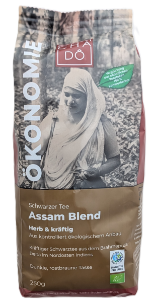 Cha Dô Ökonomie Bio Assam Blend Schwarztee-Verpackung in Braun-Grau mit Bild einer Teepflückerin im Tee-Garten. Bildbeschreibung: Standbeutel von Cha Dô mit Schwarz-Weiß-Porträt einer indischen Teepflückerin, braunem Etikett mit Aufschrift „Herb & kräftig