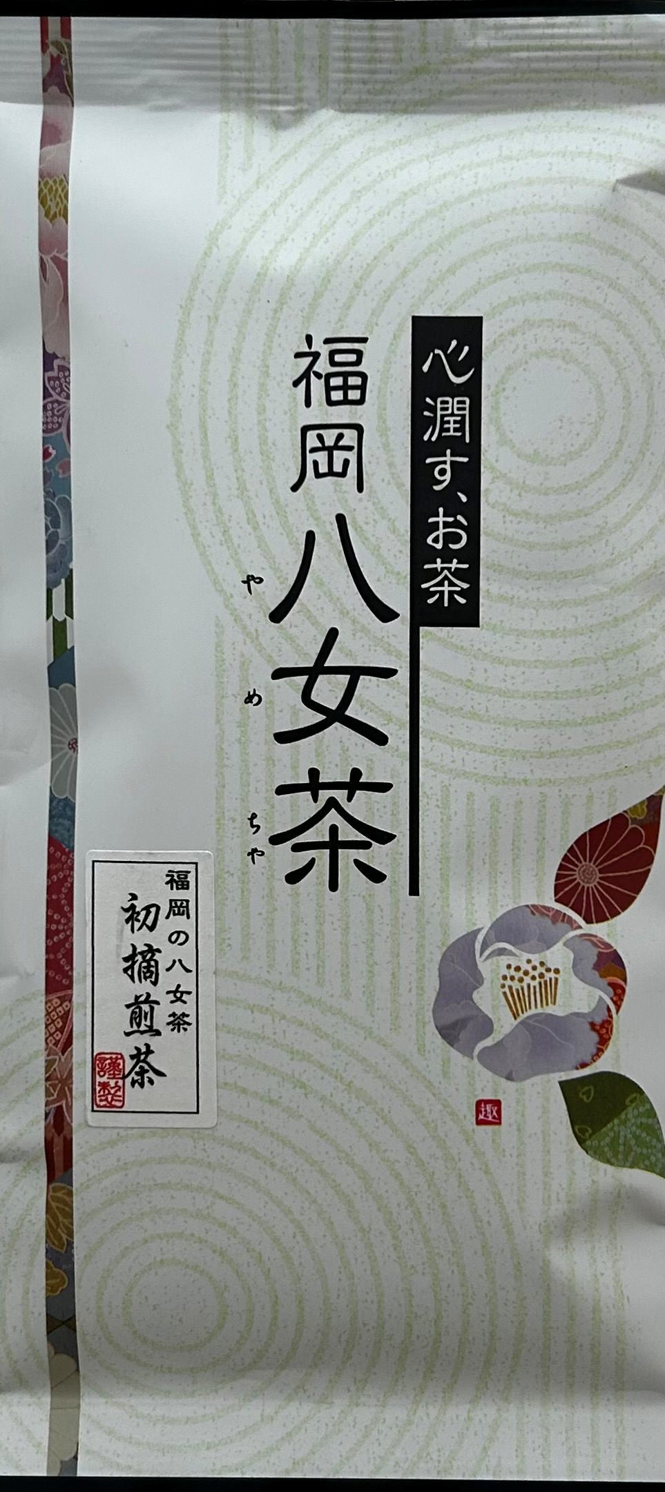 Puristische, weiße Teeverpackung, die durch ein dezentes, im Kreis geprägtes Wellenmuster im Zen-Stil besticht. Auf der rechten Seite glänzt eine kunstvoll gezeichnete, violette Kamelienblüte mit tiefgrünen Blättern. Eine feine, vertikale Linie aus schwar