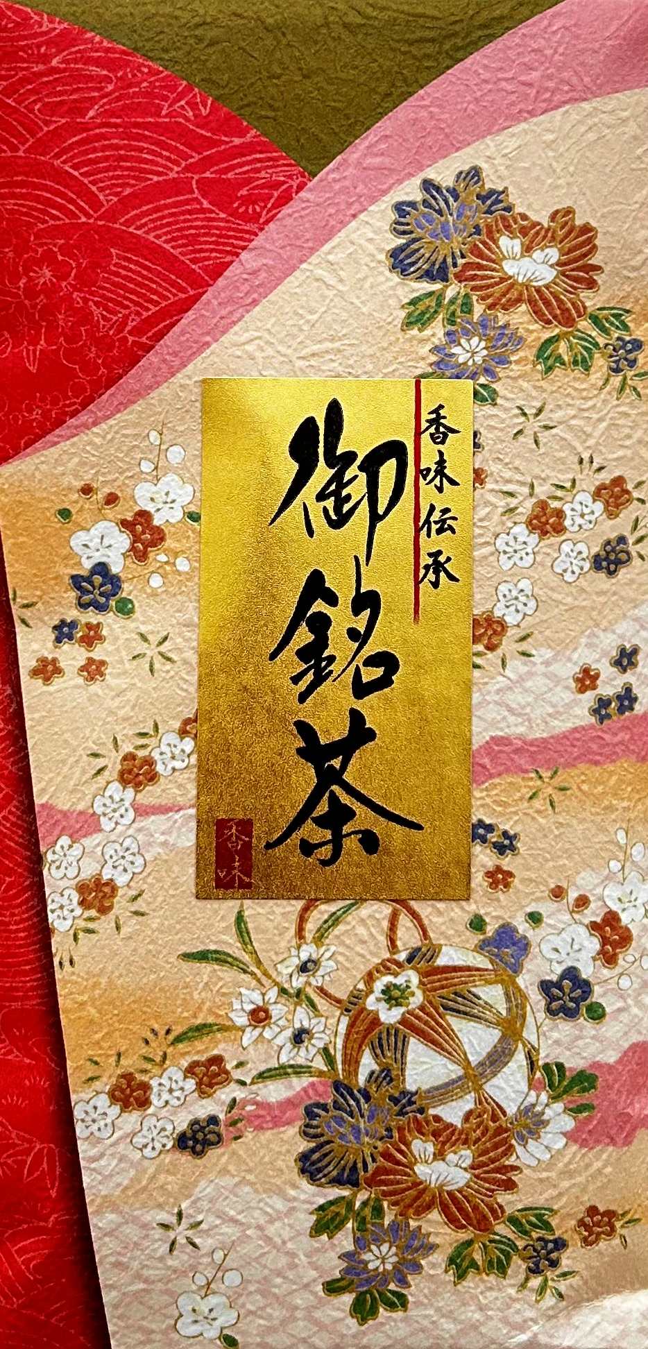 Festliche und traditionell japanische Teeverpackung im klassischen Chiyogami-Geschenkpapier-Stil. Der Hintergrund ist in einem kräftigen Zinnoberrot und glänzendem Gold gehalten, übersät mit bunten Päonien- und Chrysanthemenblüten. Mittig auf der Packung