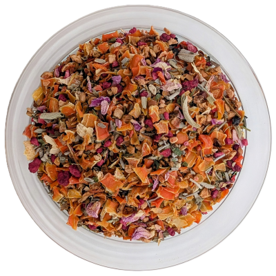 Eine runde, durchsichtige Glasschale aus der Vogelperspektive, gefüllt mit einer grob geschnittenen Teemischung. Die Optik ist stark geprägt von vielen hellorangefarbenen Karottenflocken und Apfelstücken, gemischt mit dunkelroten Hibiskusteilen, violetten