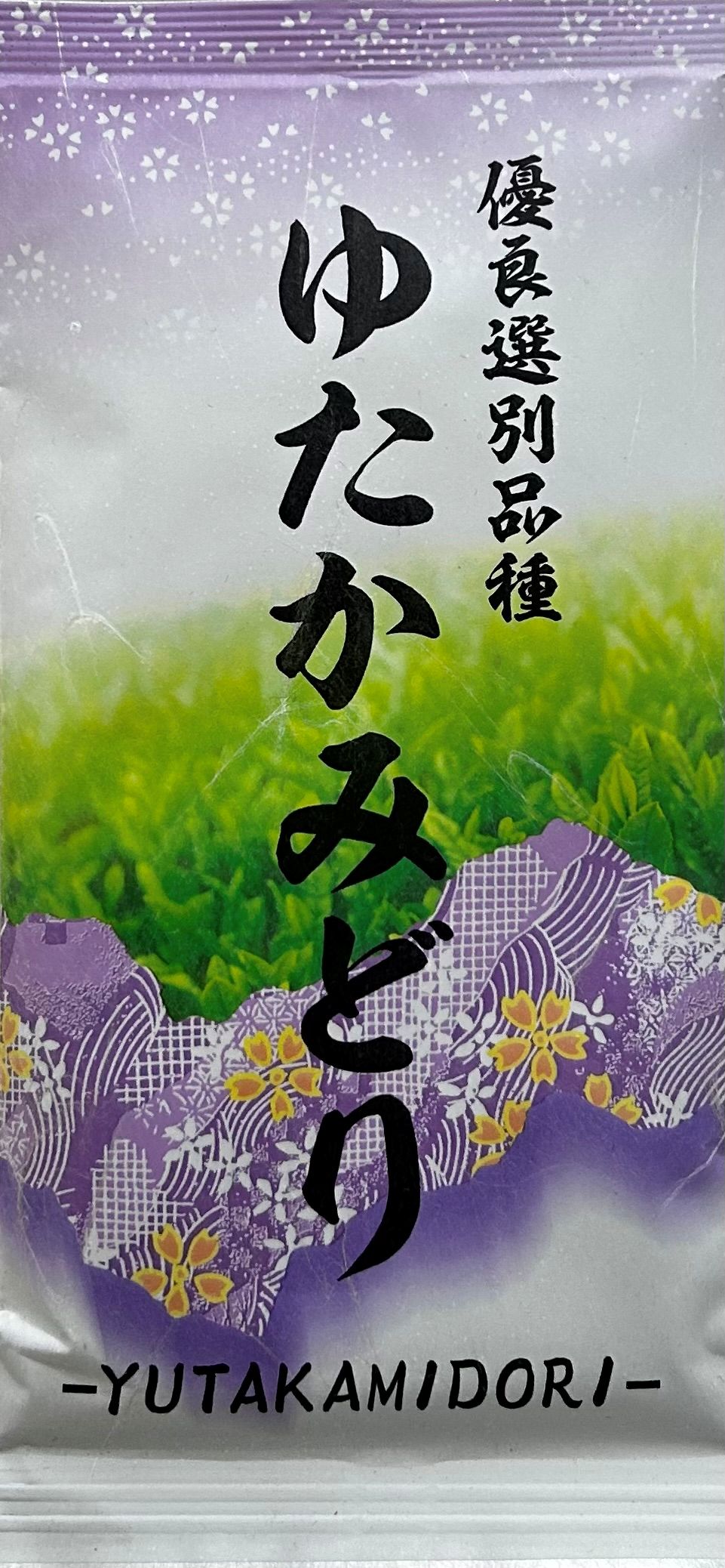 Farbenfrohe und detailreiche japanische Teeverpackung, deren Design an den edlen Stoff eines lila Seidenkimonos erinnert. Der Hintergrund ist mit goldenen und gelben Kirschblüten (Sakura) übersät, während im unteren Bereich ein leuchtend grünes Teefeld ab