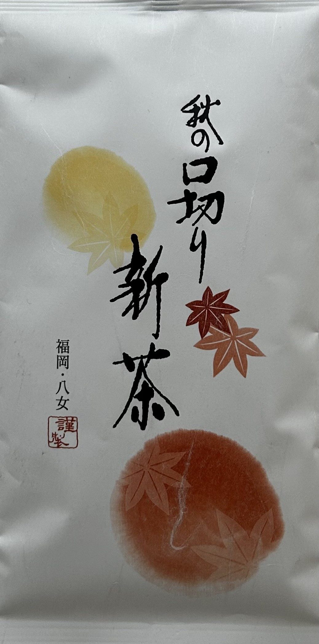 Weiße, matte Teeverpackung im herbstlichen Design. Der Hintergrund ist mit kunstvollen, runden Aquarell-Tupfern in warmen Gelb-, Orange- und Rotbrauntönen verziert, auf denen filigrane, dunkelrote Ahornblätter (Momiji) abgebildet sind. Eine vertikale Reih