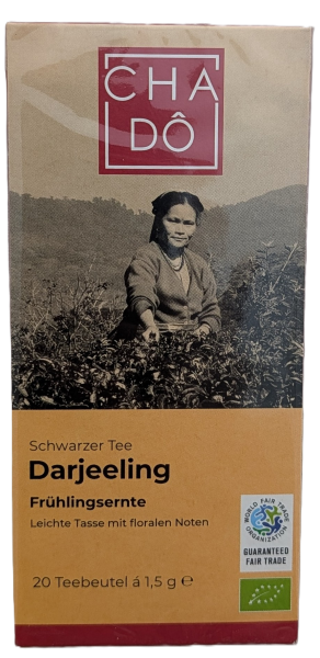 Eine quaderförmige Teeschachtel von CHA DÔ. Der obere Teil ist hellgrau mit dem roten CHA DÔ Logo und einem Schwarz-Weiß-Foto einer Teepflückerin auf einer Plantage. Der untere Teil ist orange-braun und zeigt schwarzen Text: "Schwarzer Tee Darjeeling Früh