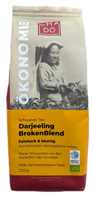 Cha Dô Ökonomie Bio Darjeeling Broken Blend Schwarztee-Verpackung in Gelb-Rot mit Bild einer Teepflückerin. Bildbeschreibung: Cremefarbene und gelbe Tee-Verpackung von Cha Dô mit historischem Schwarz-Weiß-Foto einer Teepflückerin und Bio- sowie Fairtrade-
