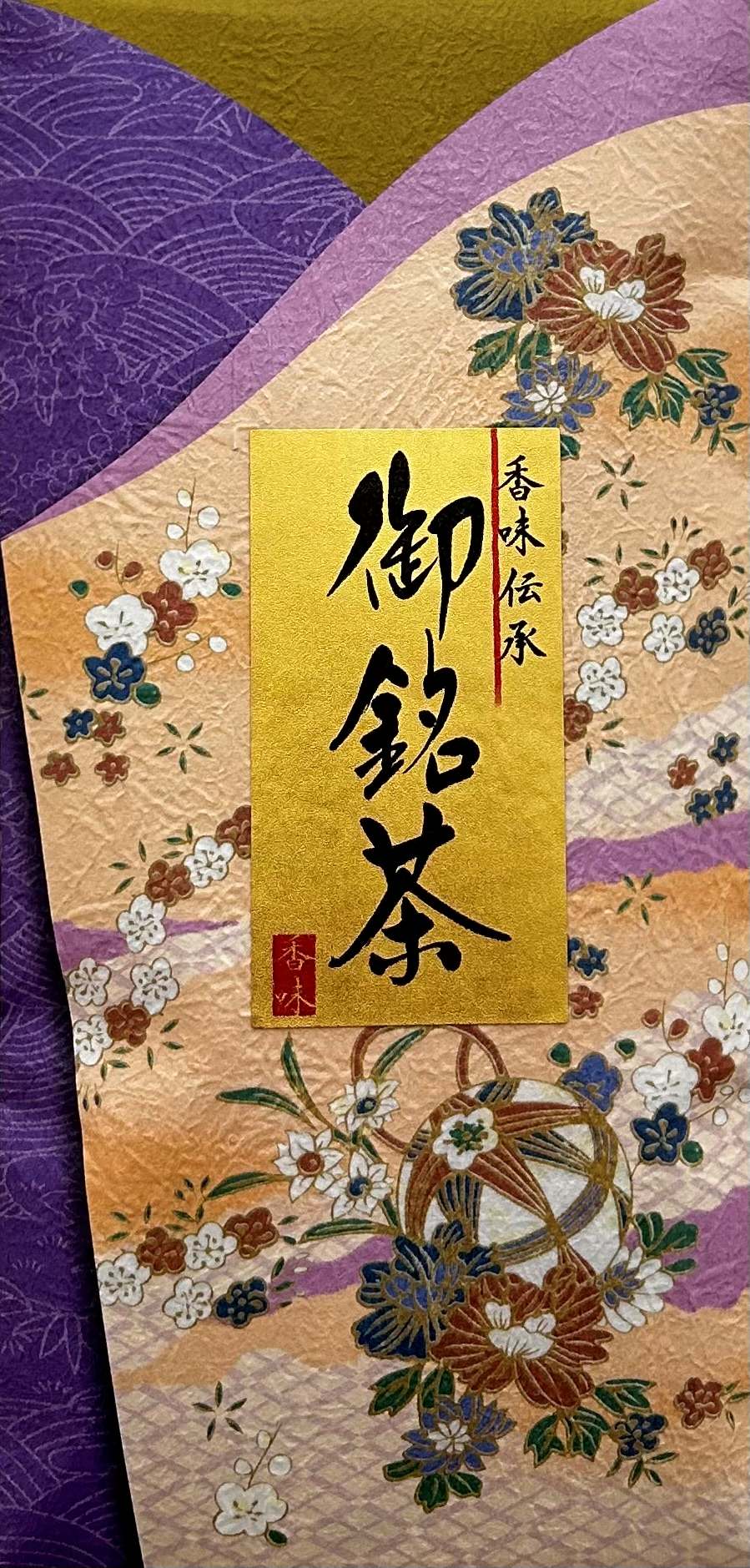 Prachtvolle japanische Geschenkverpackung für Tee, die im Hintergrund ein traditionelles Chiyogami-Muster in den Farben Gold, Violett und Dunkelblau zeigt, verziert mit bunten Blumenornamenten. Im Zentrum der Verpackung ist ein glänzendes, rechteckiges Go
